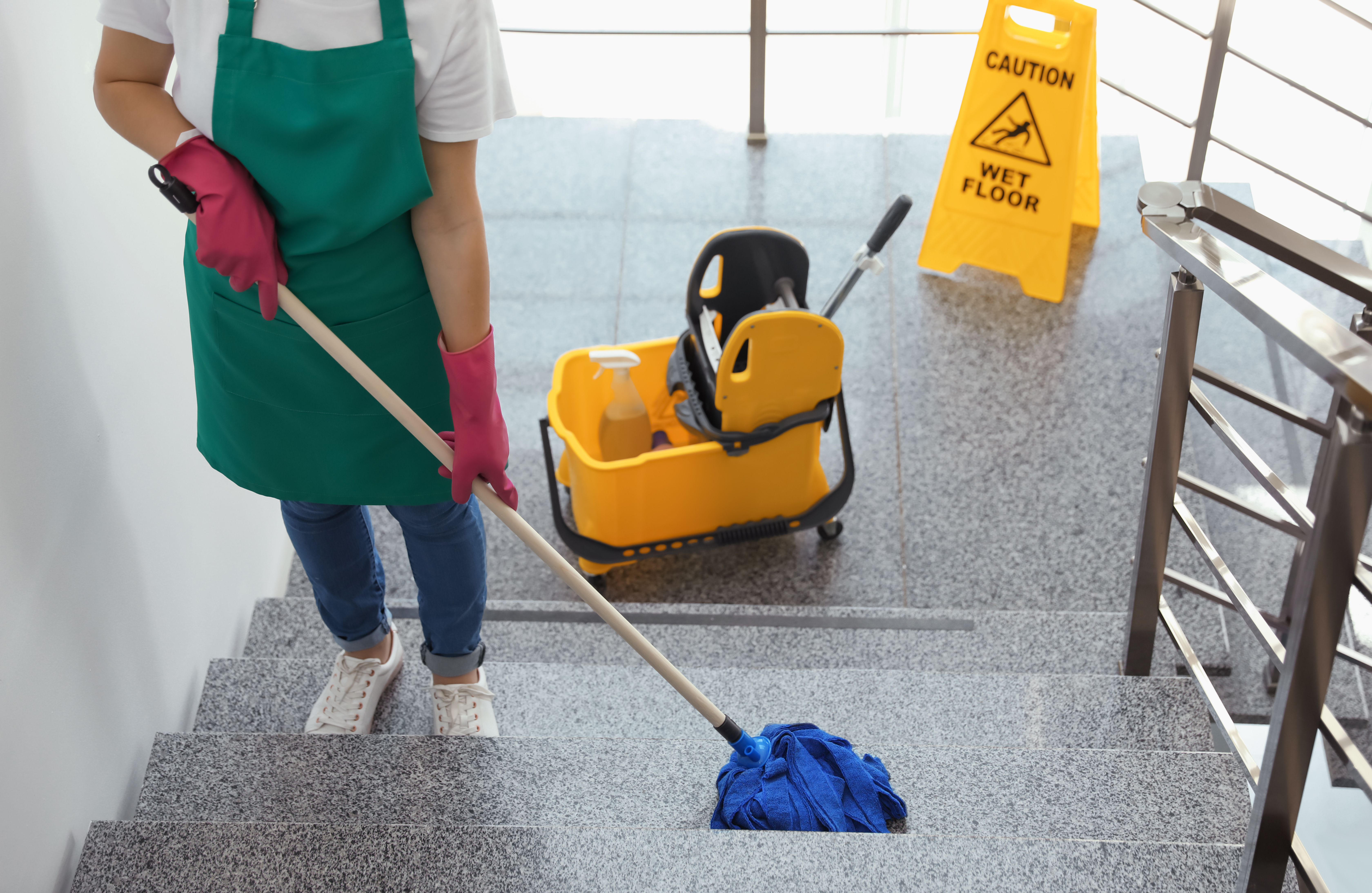 Janitor in green apron mopping stairs with wet floor caution sign and cleaning cart visible