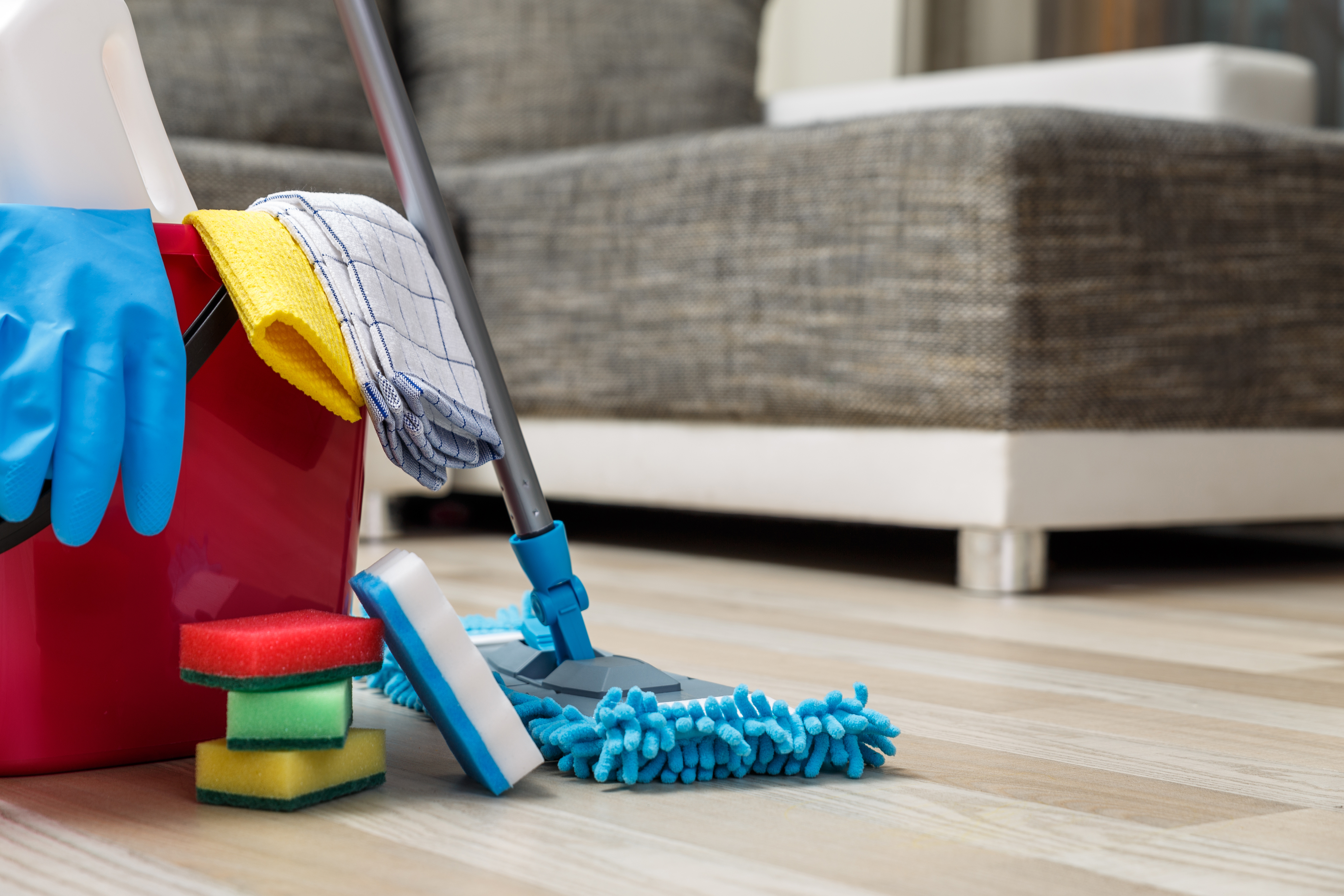 Cleaning supplies including mop, bucket, gloves, and sponges on wooden floor with sofa in background